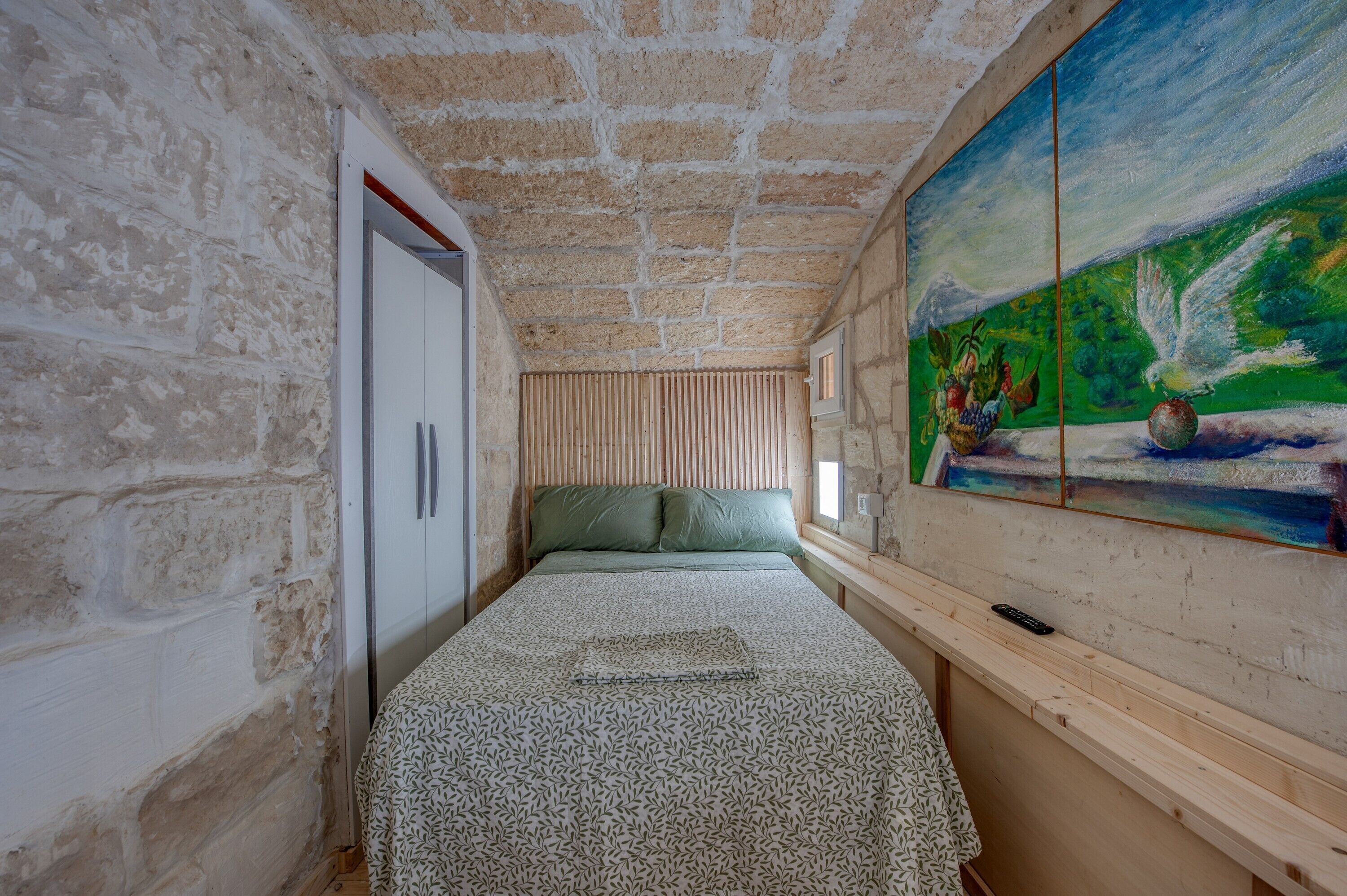Un dormitorio con pared de piedra, una cama con colcha estampada, un cuadro en la pared y una ventana con persianas.