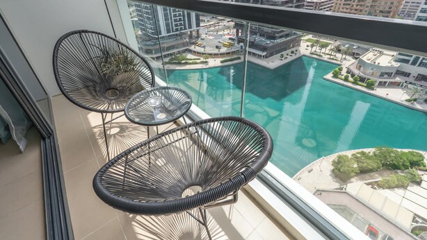 Un balcón con tres sillas de mimbre y una mesita, con vistas a una piscina y al paisaje urbano.