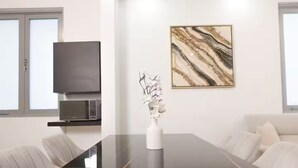 Un salon moderne avec une table à manger à plateau en verre, un téléviseur à écran plat fixé au mur, une œuvre d’art aux motifs de marbre et un canapé blanc.