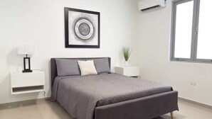 Une chambre moderne avec un lit gris, une table de chevet blanche avec une lampe, une œuvre d’art encadrée au-dessus du lit, une plante en pot et un appareil de climatisation.