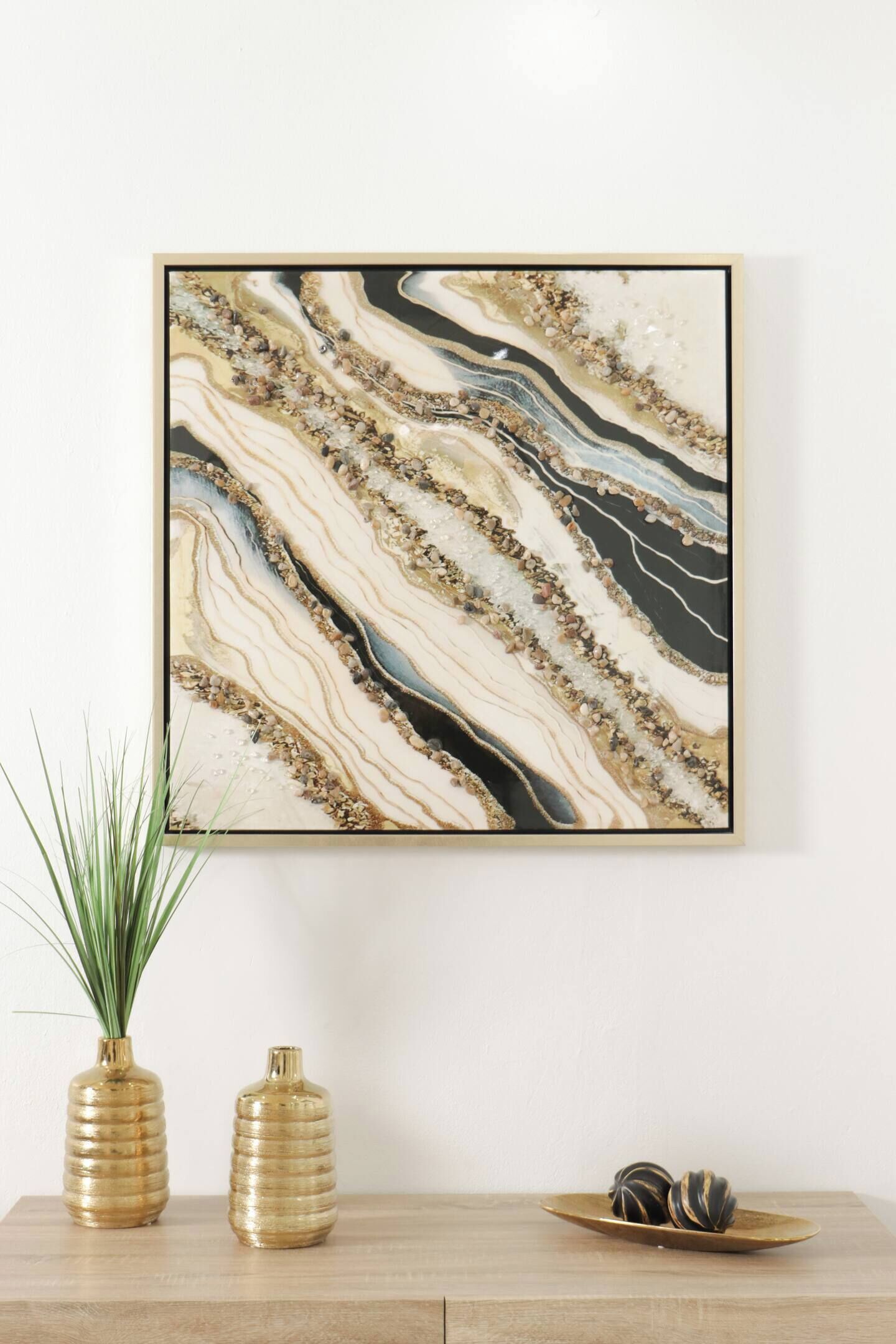 Ein gerahmtes abstraktes Gemälde mit einem marmorähnlichen Muster, zwei goldene Vasen und ein Holztablett mit dekorativen Kugeln auf einem Holztisch vor weißer Wand.