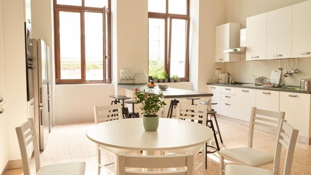 Een lichte keuken met witte kasten, een ronde eettafel en stoelen. Twee ramen zorgen voor natuurlijk licht.