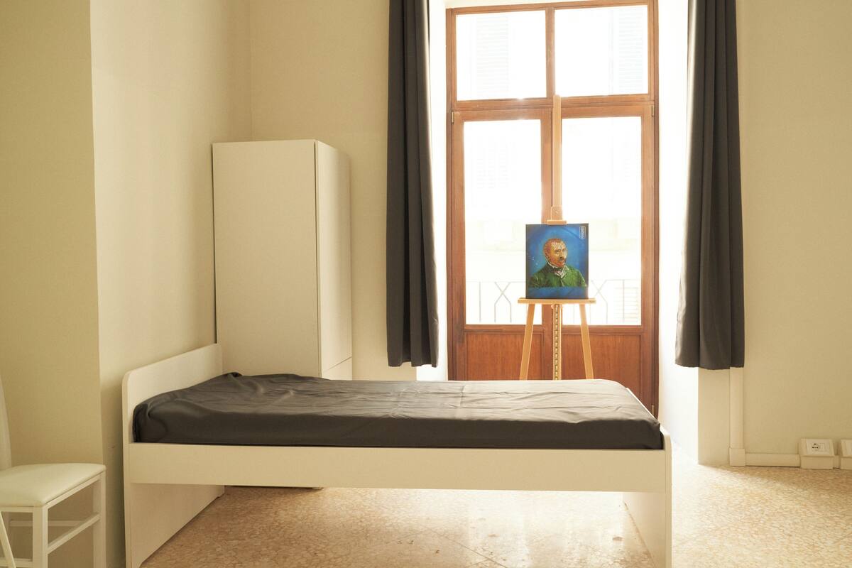 A single-bed room with a window, a small table, and a painting on an easel.