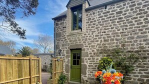 A stone building with a green door and windows, a wooden fence, and a table with chairs and a vase of flowers.