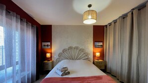 A hotel room with a large bed, two bedside lamps, a decorative headboard, and a view through sheer curtains.
