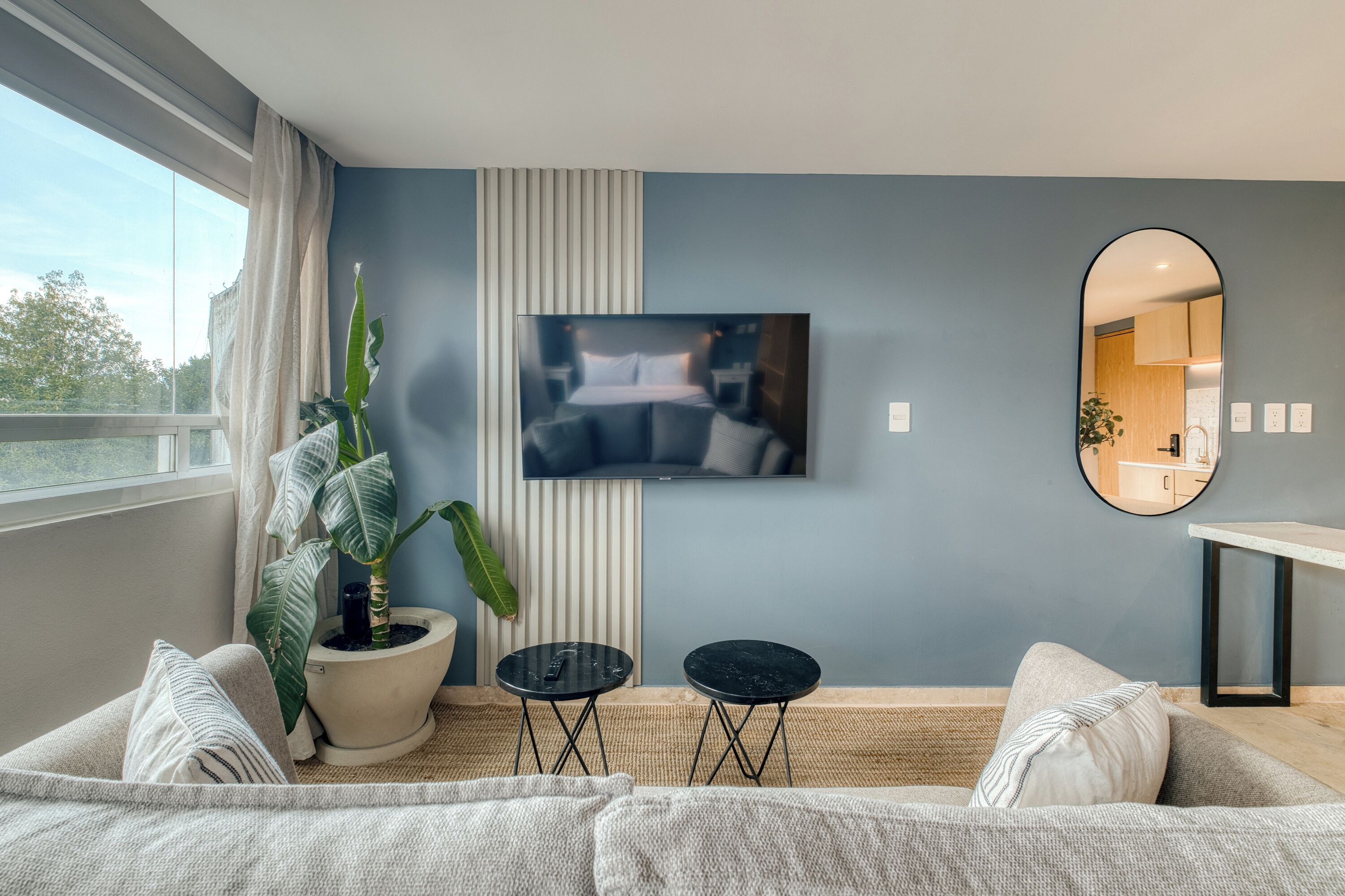 A modern living room with a large window, a flat-screen TV mounted on the wall, a round mirror, and a potted plant.