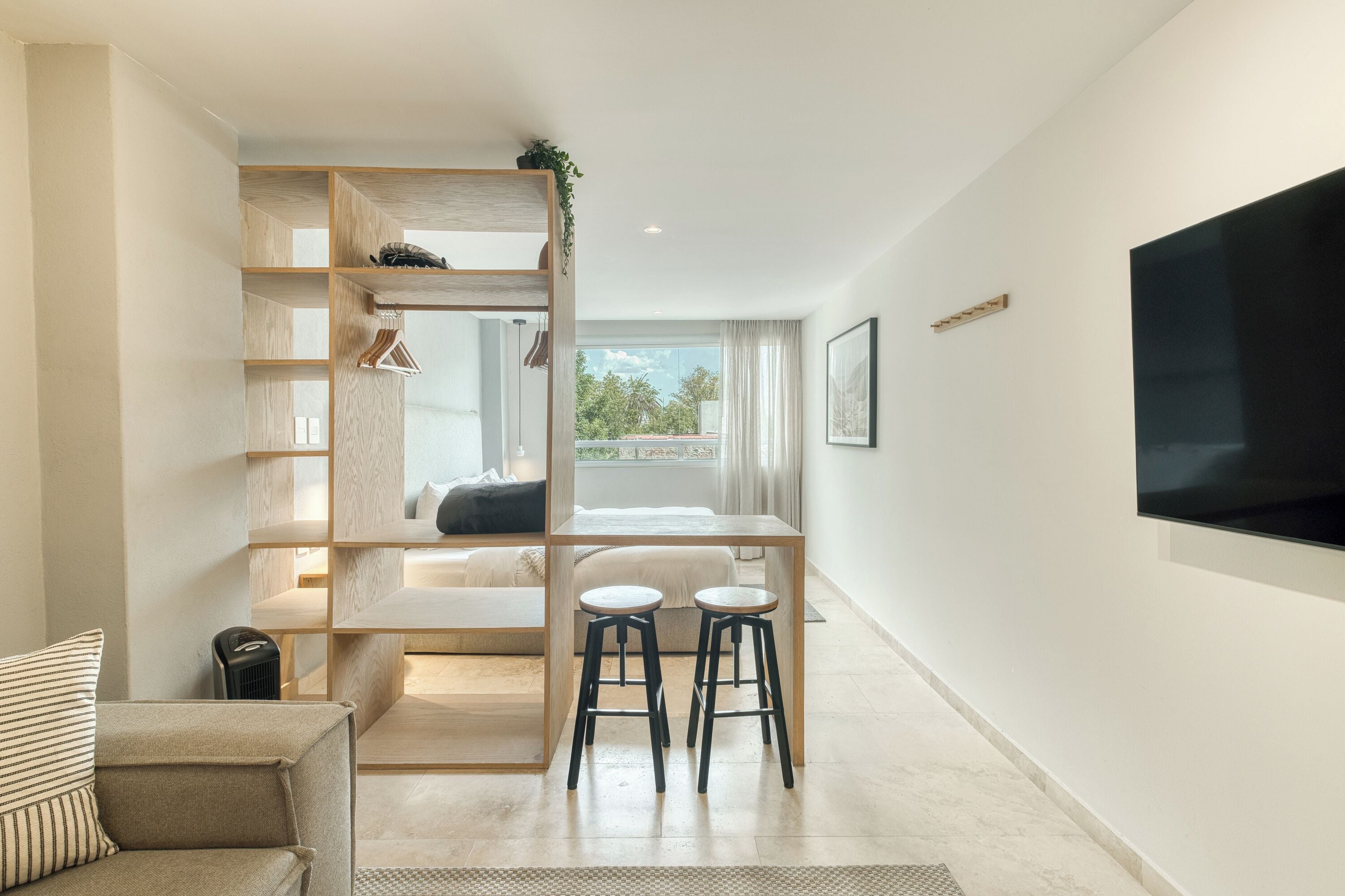 A modern, well-lit room with a built-in desk, two stools, a sofa, and a TV mounted on the wall.