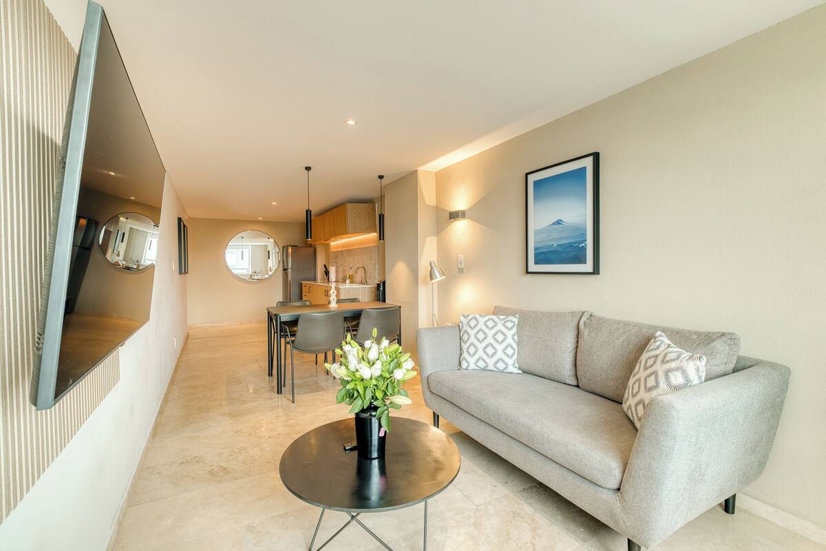 Een moderne woonkamer met een grijze bank, een ronde salontafel met een bloevas, een eethoek met een tafel en stoelen, en een keuken op de achtergrond.