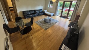 A modern living room with a black leather sofa, a glass dining table with chairs, and a small coffee table. There is a sliding glass door leading to a garden.