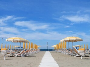 A beach with rows of sun loungers and umbrellas, a white walkway, and the ocean in the background.