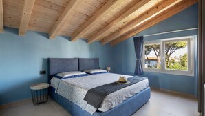 A bedroom with a wooden ceiling, a large bed with blue bedding, a bedside table, and a window with a view. - Apartment 'La Terrazza di Elisabetta' with Private Terrace, Balcony and Air Conditioning (Viareggio)