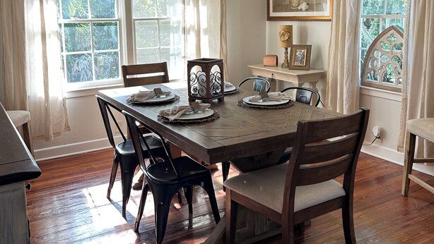 A dining room with a wooden table set for four, surrounded by chairs, a chandelier, and large windows with curtains.