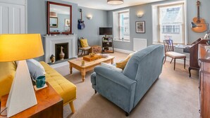 A living room with a sofa, armchair, coffee table, and a fireplace. There are guitars on the wall and a piano in the corner. - 2 Bed in Ilfracombe (oc-f31994) (Ilfracombe)