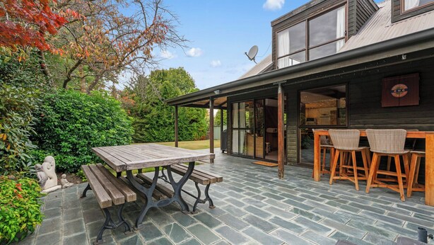 A patio with a wooden table and benches, a stone-tiled floor, and a building with large windows and a wooden deck. - So Good On Sargood - Wanaka Holiday Home (Wanaka)