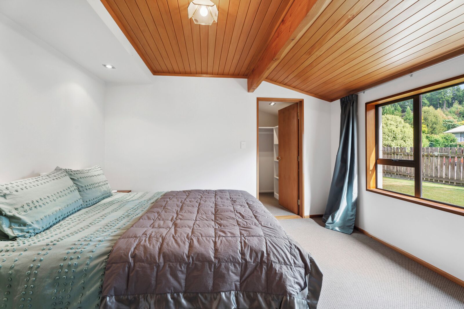 A bedroom with a large bed, a wooden ceiling, a window with a view of a backyard, and a door leading to a closet.