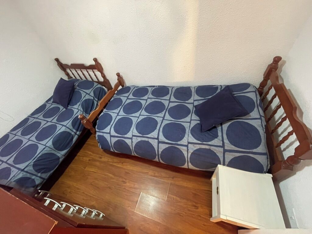 Een kleine, compacte kamer met twee houten bedden, elk voorzien van een matras en kussens met een blauw-wit patroon.