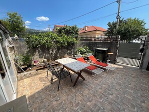 A patio area with a table and chairs, a small garden, and a view of a house in the background. - Apartment Rosa 2 (Jadranovo)