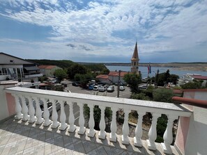 A balcony with white balustrades overlooking a parking area, a church with a tall spire, and a body of water in the distance. - Apartment Rosa 2 (Jadranovo)