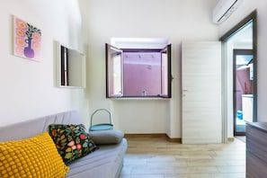 A room with a sofa, a window, a door, and a wall-mounted air conditioner.
