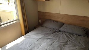 A bed with a wooden headboard, a window with curtains, and a view of the outside. - St Osyth Beach 3-bed Caravan, Sleeps 8, Parking (Clacton-on-Sea)