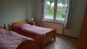 Een kamer met twee houten bedden, een nachtkastje met een lamp, een raam met gordijnen met patroon, en een radiator.