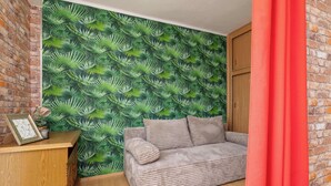 A room with a brick wall, a green leafy wallpaper, a grey sofa, and a wooden cabinet. - Cozy Studio in Katowice | Balcony and Wi-Fi (Katowice)