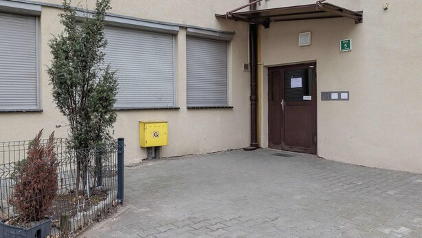 A building with a closed shuttered entrance, a yellow utility box, and a metal fence with plants. - Cozy Studio in Katowice | Balcony and Wi-Fi (Katowice)