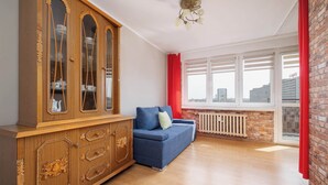 A room with a wooden cabinet, a blue sofa, a radiator, and a window with a view of buildings. - Cozy Studio in Katowice | Balcony and Wi-Fi (Katowice)