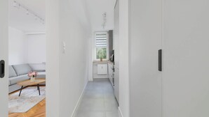 Corredor estreito com um sofá e mesa de centro à esquerda, radiador e área de cozinha no centro, e uma porta à direita.