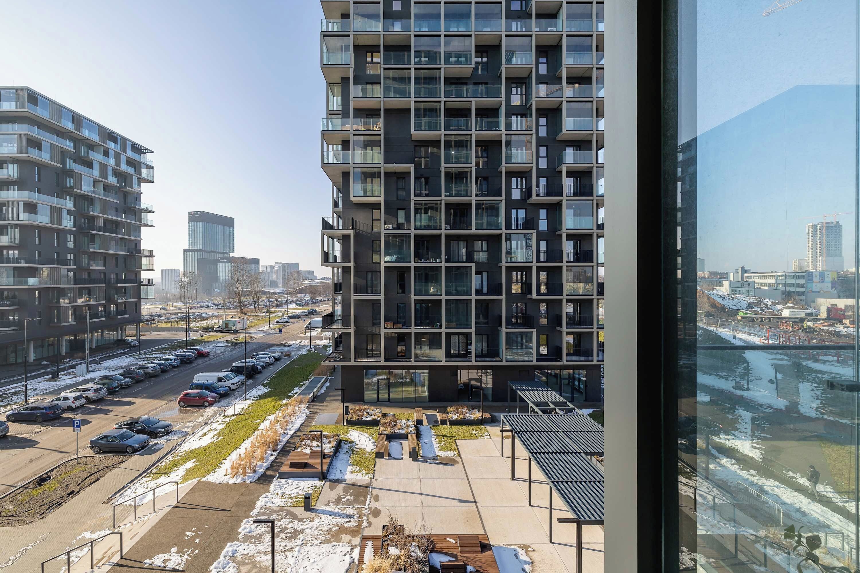 Een hoog gebouw met meerdere balkons, een parkeerplaats vol auto's en een uitzicht op een stadsbeeld.
