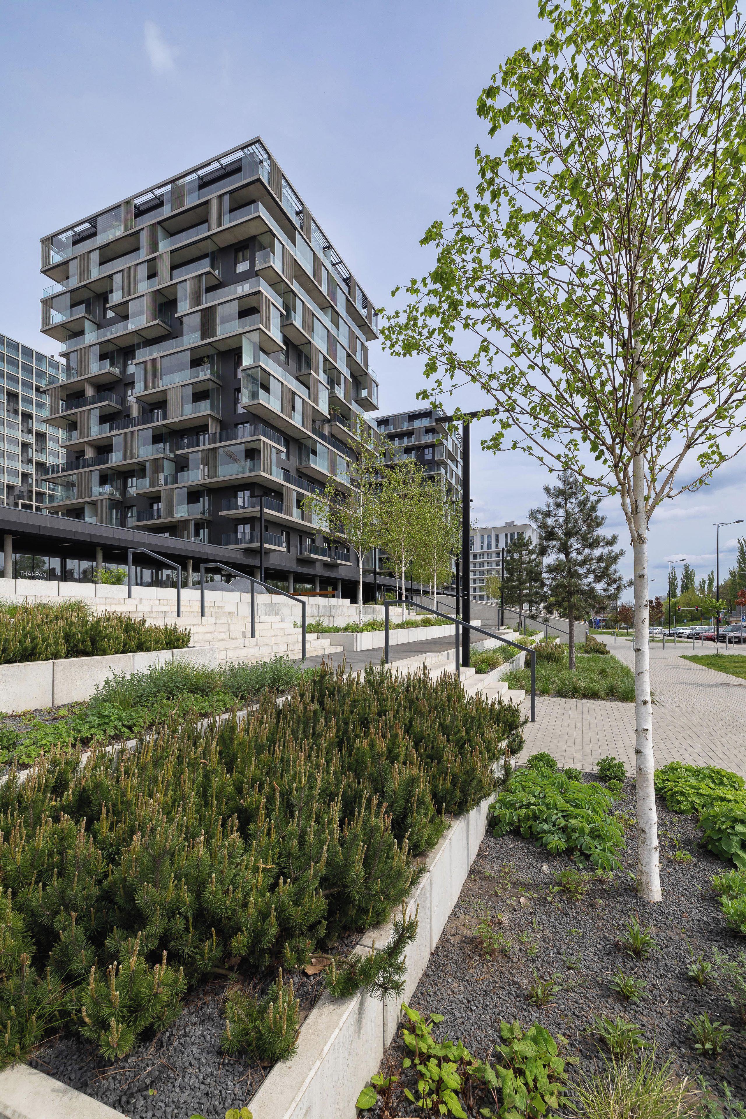 Un immeuble moderne à plusieurs étages, doté de balcons, d’un espace paysager agrémenté d’arbres et d’arbustes, et d’un chemin piétonnier.