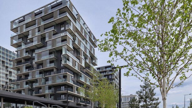 Un immeuble moderne à plusieurs étages, doté de balcons, d’un espace paysager agrémenté d’arbres et d’arbustes, et d’un chemin piétonnier.
