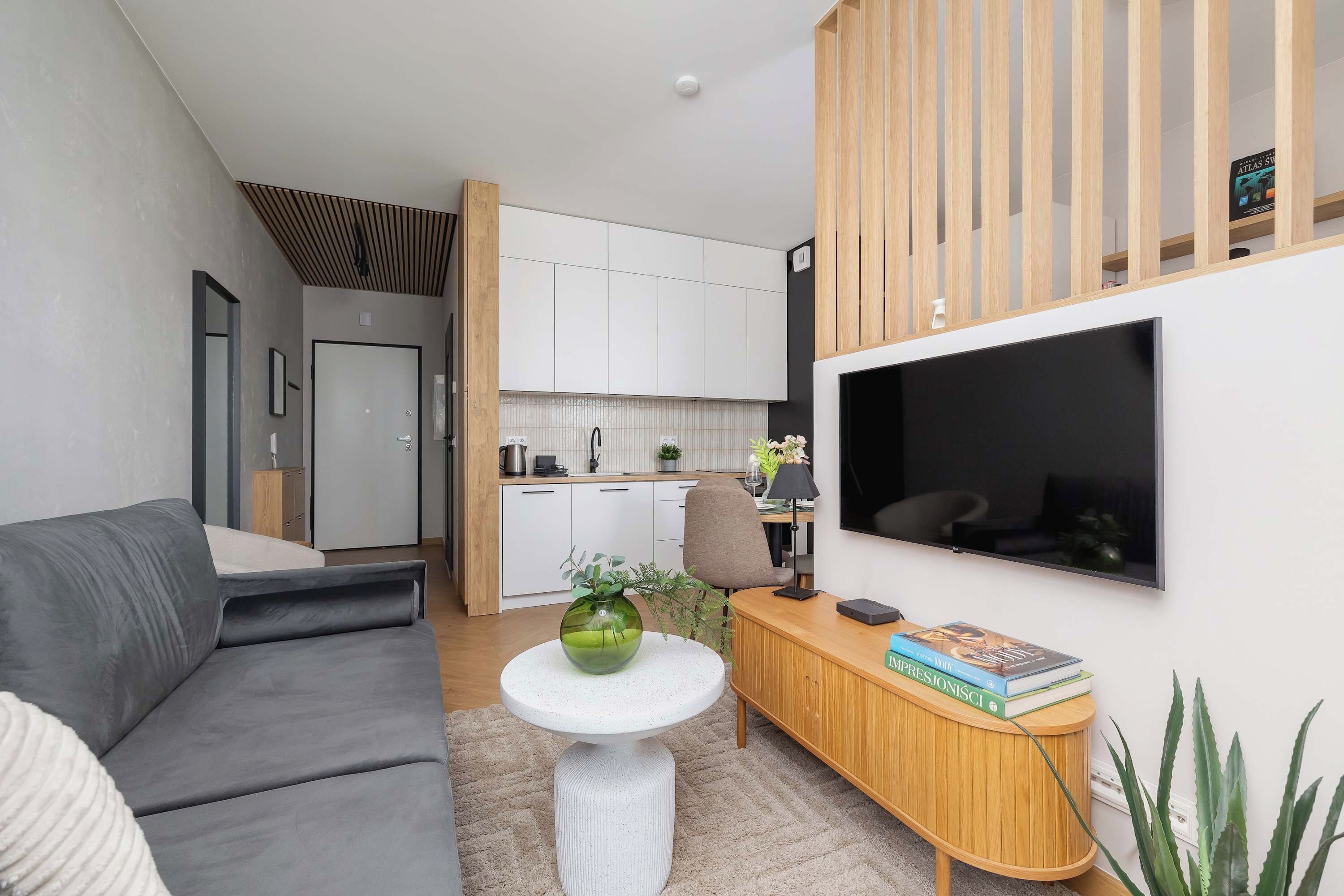 Een moderne woonkamer met een grijze bank, een ronde witte tafel met een groene vaas, een houten console met boeken en een aan de muur bevestigde televisie.