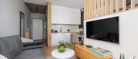 A modern living room with a grey sofa, a round white table with a green vase, a wooden console table with books, and a TV mounted on the wall.
