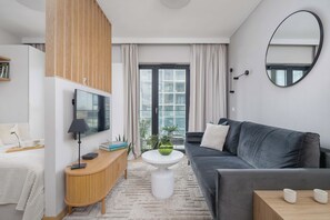 Un salon moderne avec un canapé gris, une table basse blanche et un téléviseur fixé au mur. Un panneau en bois orne le mur, et une grande fenêtre est agrémentée de rideaux.