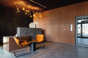 A modern reception area with a reception desk, a seating area with a chair, a wall-mounted clock, and a ceiling with hanging lights.