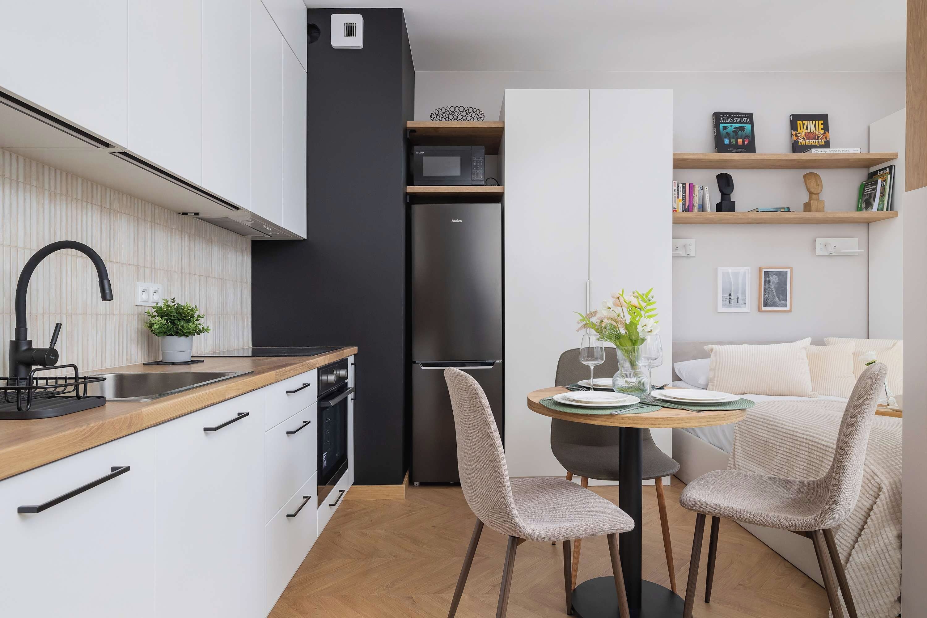 Eine moderne Küche mit weißen Schränken, einem schwarzen Kühlschrank und einer Holzarbeitsplatte. Es gibt eine Essecke mit zwei Stühlen und einem Tisch.