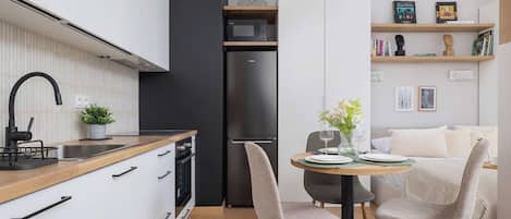 Una cocina moderna con armarios blancos, un refrigerador negro y una encimera de madera. Hay una zona de comedor con dos sillas y una mesa.