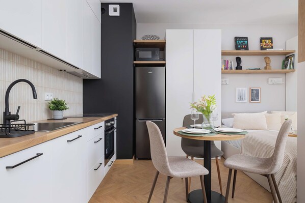 A modern kitchen with white cabinets, a black refrigerator, and a wooden countertop. There is a dining area with two chairs and a table.
