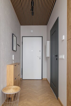 Un couloir doté d’un plancher en bois, une porte avec une poignée, une petite table ronde dont le plateau est fait de fibres tressées, et un luminaire fixé au mur.