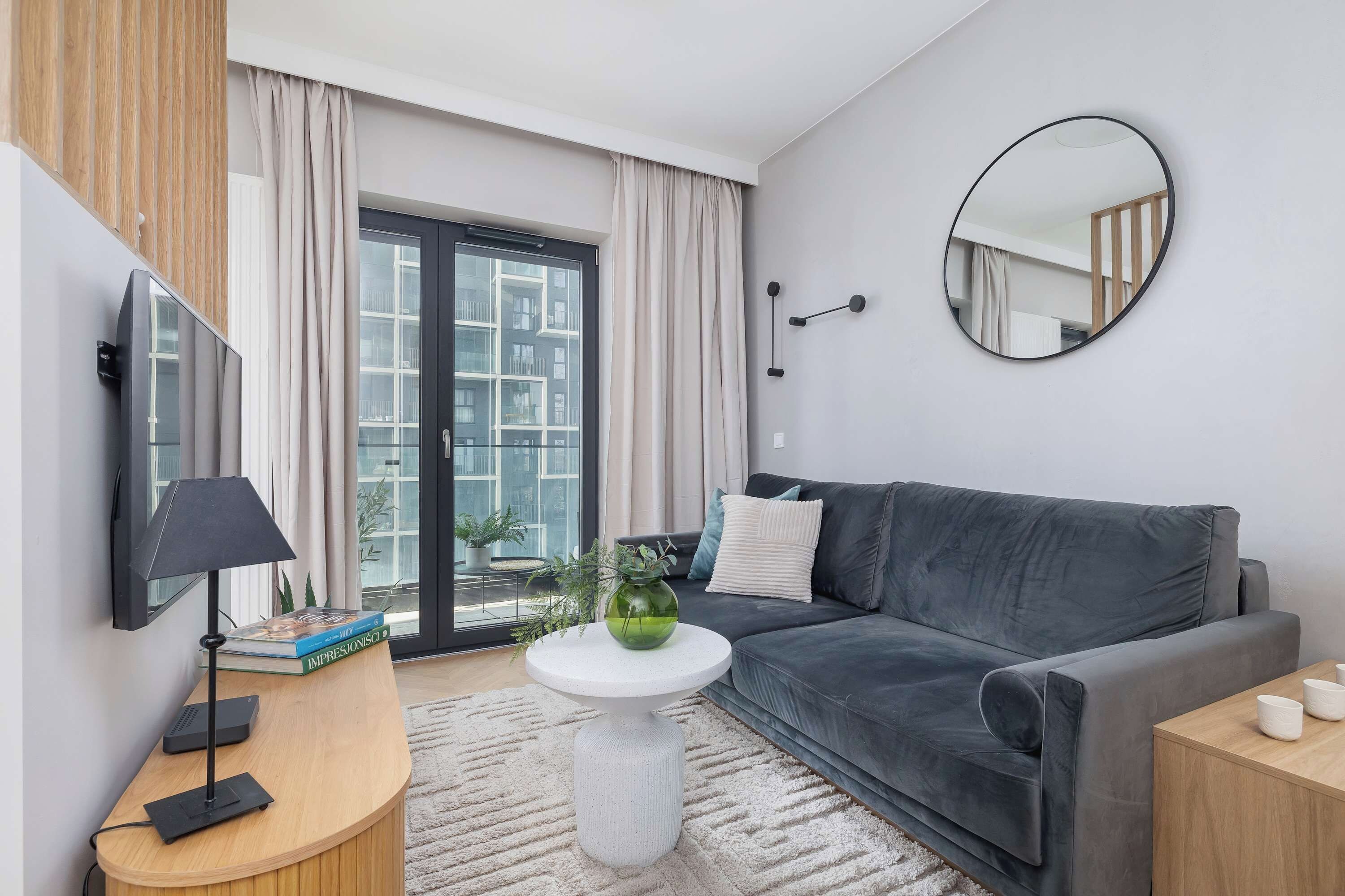 Un salon moderne avec un canapé gris, un miroir rond, une petite table blanche ornée d’un vase vert et un téléviseur posé sur un meuble en bois.