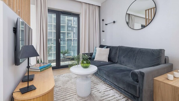 Een moderne woonkamer met een grijze bank, een ronde spiegel, een klein wit tafeltje met een groene vaas, en een tv op een houten dressoir.