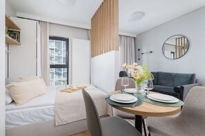 Ein modernes Hotelzimmer mit Bett, Essbereich mit Tisch für zwei Personen, Sofa und großem Fenster.