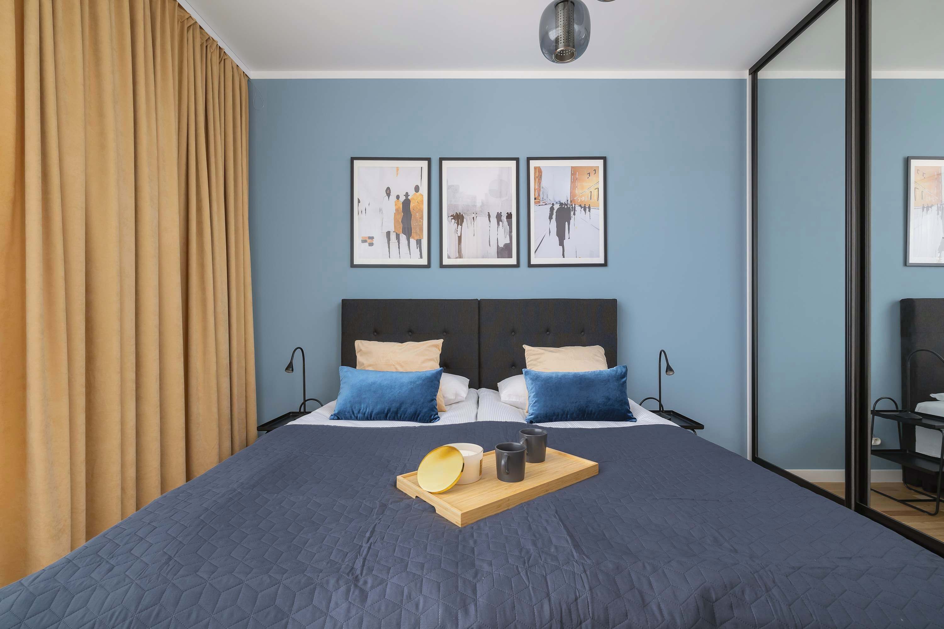 A modern hotel room with a large bed, two side tables, and a bedside table with a lamp. There are three framed artworks on the wall above the bed.