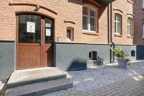 A brick building with a wooden door, a sign indicating a room number, and a potted plant in the foreground.