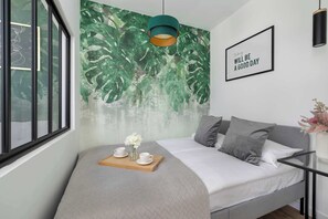 Une chambre à coucher moderne avec une grande fenêtre, un lit avec une literie grise, une table de chevet en bois avec des tasses et des fleurs, une fresque murale verte et une citation inspirante sur le mur.