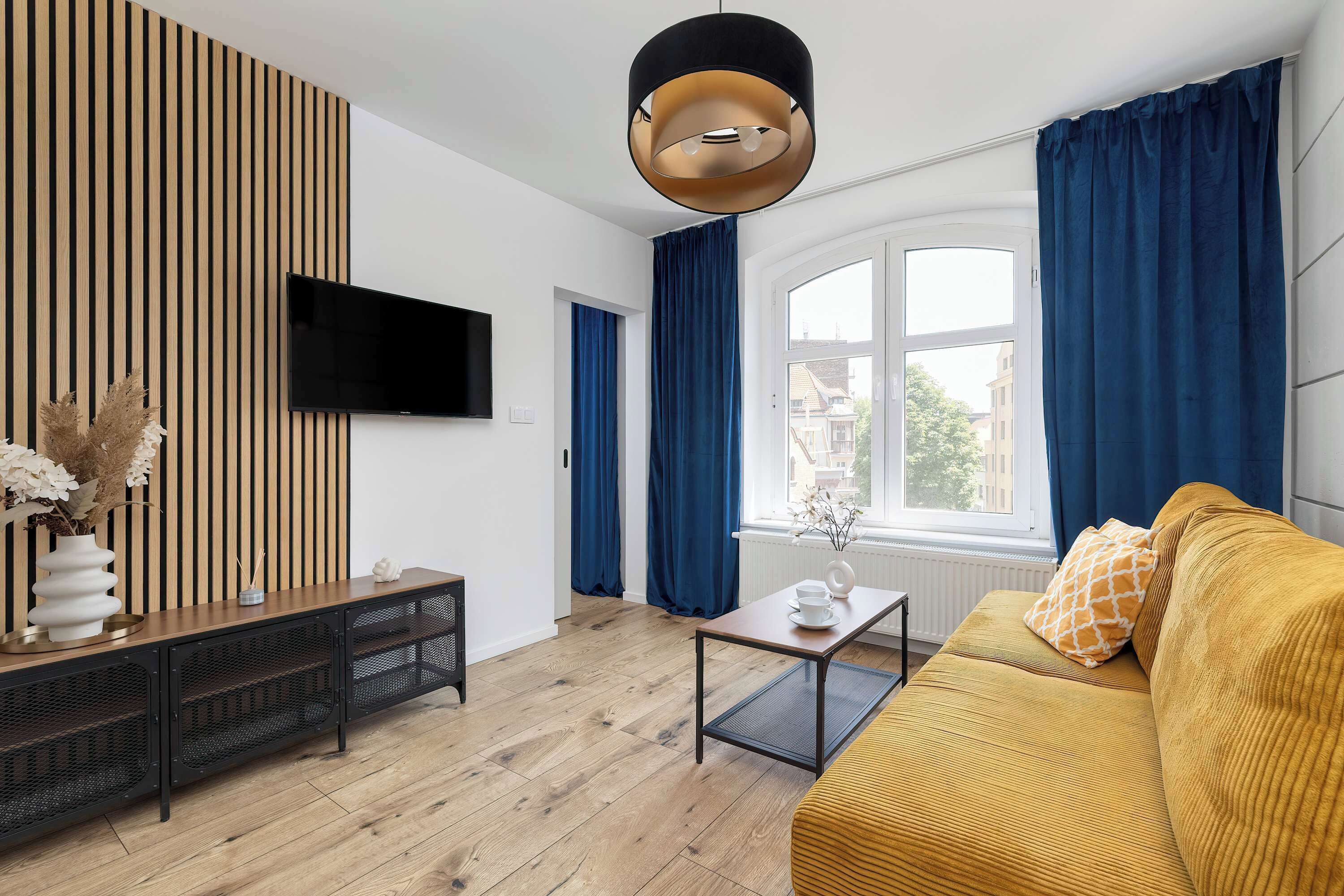 A modern living room with a wooden panel wall, a flat-screen TV, a sofa, a coffee table, and a window with curtains.