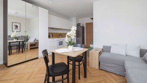 Een moderne woonkamer met een grijze bank, een houten eettafel met twee stoelen, een keuken met kastjes en een koelkast, en een grote spiegel die de kamer reflecteert.