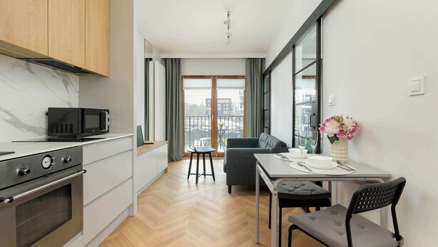 Een moderne keuken met een magnetron, oven en houten kastjes. Er is een zithoek met een tafel, stoelen en een bloevas.