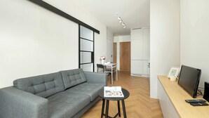Een moderne woonkamer met een grijze bank, een klein rond tafeltje met een tijdschrift, en een televisie op een houten bureau.